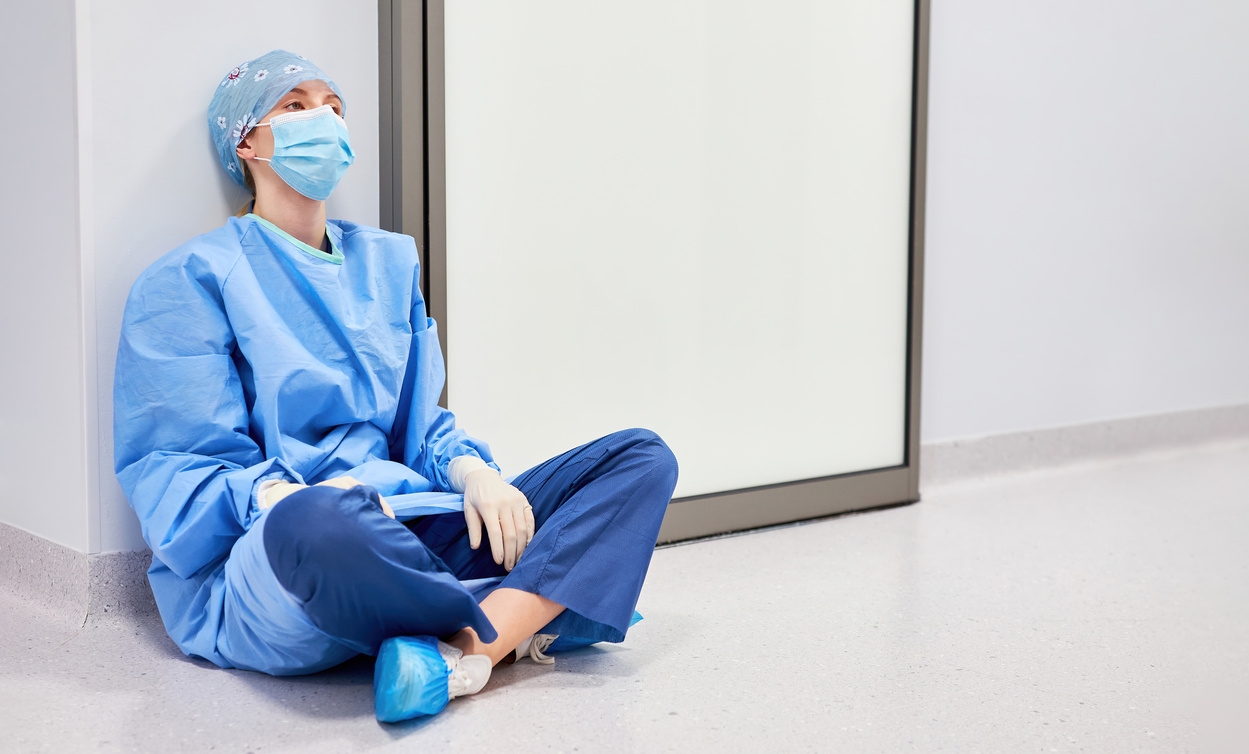 A health care worker in scrubs and a mask sits exhausted on a hospital corridor floor